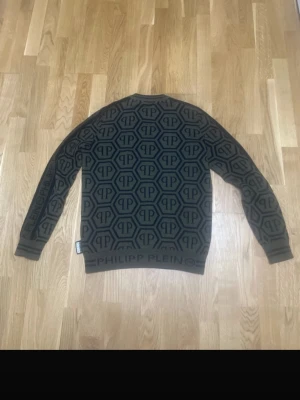 Mönstrad tröja från Philipp Plein - Snygg mörkgrön tröja från Philipp Plein med svart grafiskt logomönster över hela plagget. Tröjan har rund hals, ribbade muddar och tydlig logga på nederkanten. Perfekt för dig som gillar exklusiv streetstyle och vill sticka ut.