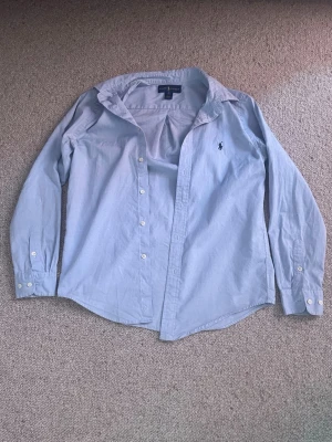 Ljusblå skjorta från Ralph Lauren - Klassisk ljusblå skjorta från Ralph Lauren med broderad logga på bröstet. Skjortan har knappar framtill, krage och långa ärmar som kan vikas upp och fästas med knapp. Tillverkad i mjuk bomull för en skön känsla. Pris kan diskuteras såklart!