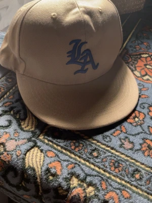 Beige LA-keps med blå logga - Säljer en beige keps med blå LA-logga broderad framtill. Klassisk rak skärm och justerbar baksida. Tillverkad i slitstarkt tyg, perfekt för dig som gillar streetwear och sportig stil.