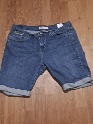 Blå jeansshorts från Levi's - Klassiska blå jeansshorts från Levi's med upprullade benslut Shortsen har knappgylf, bälteshällor och dekorativa sömmar på bakfickorna. 
