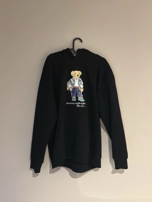 Svart hoodie Polo Bear Ralph Lauren - Svart hoodie från Ralph Lauren med ikoniskt Polo Bear-tryck på bröstet. Klassisk känguruficka framtill och huva. Perfekt för dig som vill ha en stilren men lekfull look. Mjuk och skön bomullskvalitet.