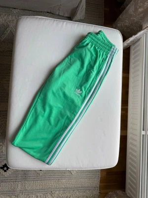 Gröna Adidas träningsbyxor - Säljer ett par fräscha gröna mjukisbyxor från Adidas med klassiska vita och lila ränder längs sidorna. Byxorna har resår i midjan och Adidas-logga på låret. Perfekta för chill eller träning, riktigt snygga och bekväma.