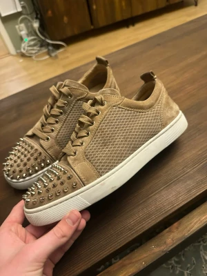 Beige Christian Louboutin sneakers - Snygga beige sneakers från Christian Louboutin med meshpaneler och mockadetaljer. Skorna har coola silverfärgade nitar på tån och den klassiska röda sulan. Perfekta för dig som vill sticka ut med din stil.