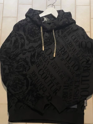 Versace Jeans Couture Hoodie – Svart - Snygg och exklusiv hoodie från Versace Jeans Couture i svart ton-i-ton barockmönster. Stilren men samtidigt iögonfallande design med diskret logotryck i tyget och guldfärgade kedjedetaljer i huvan som ger en lyxig känsla. Mjuk och skön kvalitet, perfekt både till vardags och för en mer uppklädd streetwear-look.  