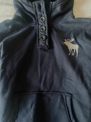 Mörkblå hoodie med älgtryck - En jättefin, unik mörkblå hoodie från Abercrombie & Fitch som inte säljs längre. Den är medium men passar mig som vanligtvis bär small.  Den är i 100% bomull och är bekväm att ha på sig. Passar till alla tillfällen. Inga synliga defekter. Katt finns i hemmet, men tvättar innan.