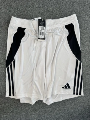 Vita sportiga shorts från Adidas - Snygga vita shorts från Adidas med svarta detaljer och klassiska ränder på sidorna. De har elastisk midja med snörning och är tillverkade i ett lätt och svalt polyestermaterial. Perfekta för fotboll eller träning.