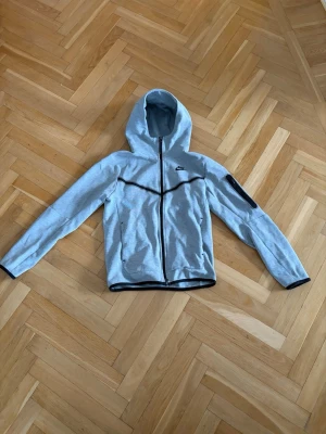 Nike Tech Fleece grå - Säljer denna tech fleece från Nike med hel dragkedja framtill och svart kant vid ärmslut och nederkant. Tröjan har huva och en ficka med dragkedja på ena ärmen. Perfekt för en sportig och avslappnad stil.