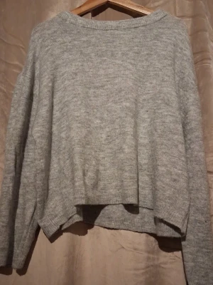 Grå oversize stickad tröja - Mysig grå stickad tröja i oversize-modell med rund halsringning och långa ärmar. Tröjan har en något kortare och lös passform, perfekt för lager på lager. Materialet ser ut att vara mjukt och skönt, troligen en ull- eller akrylblandning.