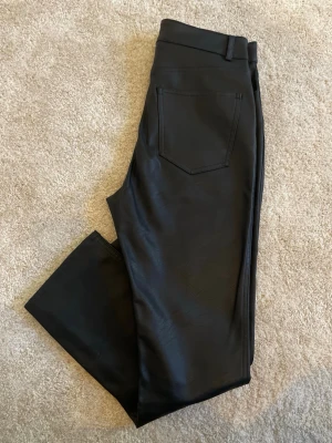 Svarta skinnbyxor med raka ben - Snygga svarta byxor med coated finish som ger en läderliknande look. Raka ben, klassisk femficksmodell och bälteshällor. Perfekta för dig som vill ha en edgy stil och gillar trendiga detaljer.