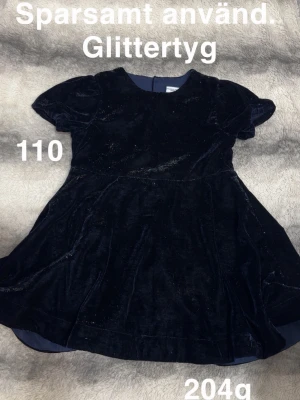 Mörkblå glittrig klänning barn - Söt mörkblå klänning i glittrigt tyg med korta puffärmar och rund hals. Kjolen har en lätt utställd form och tyget ger en lyxig känsla. Perfekt för speciella tillfällen när man vill glänsa lite extra.🌸 Sparsamt använd. Glittertyg. Storlek 110. 