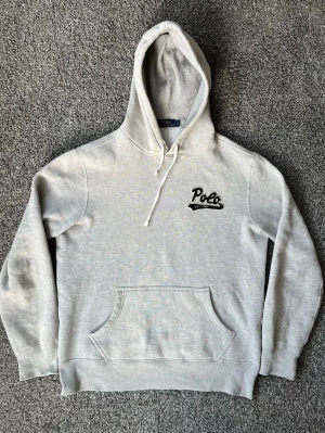 Grå hoodie från Polo Ralph Lauren - Kolla in mina andra plagg!!!  Snygg ljusgrå hoodie från Polo Ralph Lauren med broderad svart Polo-logga och text 'EST. 1967' på bröstet. Klassisk känguruficka framtill, dragsko i huvan och mjukt bomullsmaterial. Perfekt för en avslappnad och stilren look.
