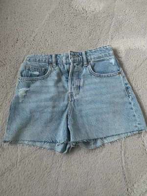 Ljusblå denimshorts - Säljer ett par ljusblå denimshorts med hög midja och rå, fransig kant. Shortsen har klassisk femficksdesign och slitna detaljer framtill för en avslappnad look. Perfekta till sommaren och lätta att matcha med olika stilar.