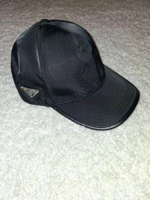 Svart keps från Prada i nylon - Snygg svart keps från Prada i glansigt nylonmaterial med klassisk böjd skärm och ikonisk metallogo på sidan. Perfekt accessoar för en clean och stilren look.