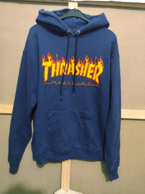 Blå Thrasher hoodie med flames - Snygg blå hoodie från Thrasher med ikoniskt flames-tryck i gult och orange på bröstet. Klassisk känguruficka framtill, huva med dragsko och mjukt material. Perfekt för dig som gillar streetwear och skatekultur.