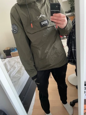 Olivgrön vindjacka från Dope - Snygg olivgrön vindjacka från Dope med stor magficka, huva och svarta samt orange detaljer. Jackan har patch på ärmen och logga på bröstet. Perfekt för dig som vill ha en stilren och funktionell jacka till Skidor. Heöt oandvänd, Glid på med prisförslag
