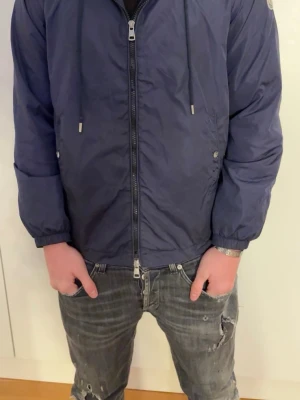 Marinblå Moncler vindjacka med dragkedja - Snygg marinblå vindjacka med hel dragkedja framtill och justerbar huva med snören. Jackan har två sidofickor med tryckknappar och resår vid ärmslut och midja. Perfekt för blåsiga dagar och enkel att matcha till olika outfits.