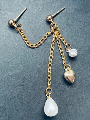 By prisförslag- 1 st dubbel Guldörhänge med kedjor och berlocker - Unika örhänge i guldfärgad metall med dubbla kedjor och tre berlocker: ett hjärta, en droppformad pärla och en gnistrande sten. Snyggt statement-smycke som ger en trendig och lekfull look. Perfekt för dig som gillar detaljer och mixade material.