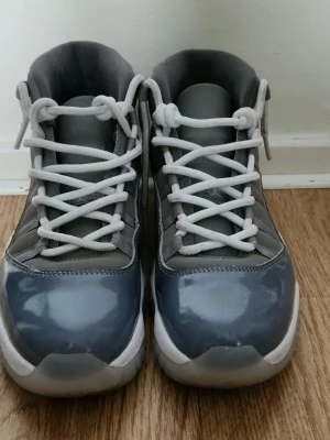 Grå Air Jordan 11 sneakers - Snygga Air Jordan 11 sneakers i cool grå färg med vita detaljer och vita skosnören. Skorna har en blank finish på tån och sidorna, samt den klassiska Jumpman-loggan på hälen. Ovandelen är i textil och syntet, sulan är vit och genomskinlig.