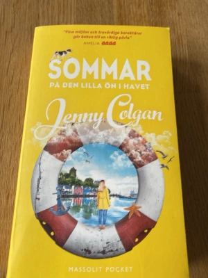Sommar på den lilla ön i havet - En färgstark pocketbok av Jenny Colgan med gul framsida och illustration av en livboj och havsmotiv. Boken handlar om Flora som återvänder till en liten skotsk ö och möter både gamla minnen och nya utmaningar. Perfekt för dig som gillar romantisk och varm feelgood.