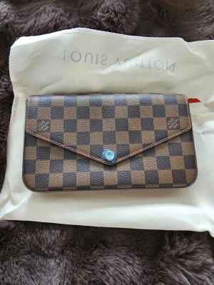 Damier kuvertväska - Snygg kuvertväska i klassiskt brun färg.. Väskan har guldfärgad kedja, röd insida och matchande accessoarer som korthållare och dragkedjeförsett fack. Tillverkad i canvas med FU läderdetaljer och tryckknapp framtill.