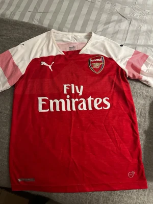 Arsenal matchtröja Puma röd/vit - Snygg Arsenal matchtröja från Puma i rött och vitt med klubbmärke på bröstet och Fly Emirates-tryck. Tröjan har korta ärmar med röda detaljer och Puma-logga på ärmen. Perfekt för fotbollsfans som vill visa sitt stöd på matchen eller träningen.