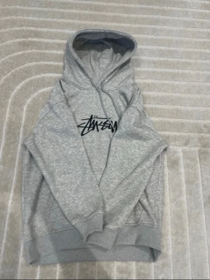 Grå Stüssy hoodie med logga - Säljer en grå hoodie från Stüssy med den klassiska svarta loggan tryckt på bröstet. Hoodien har huva med dragsko, magficka och ribbade muddar vid ärmar och nederkant. Tillverkad i mjukt bomullsmaterial som är skönt att ha på sig.