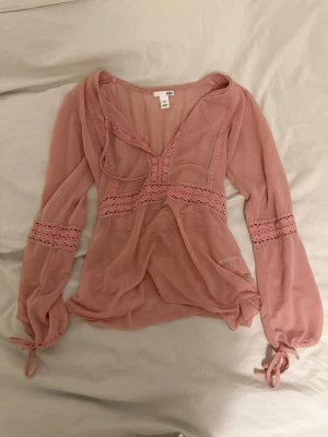 Rosa topp med spetsdetaljer - Säljer eb rosa omlottopp från H&M med v-ringning och spetsdetaljer. Toppen har långa ärmar med knyt vid handlederna och är tillverkad i ett tunt, genomskinligt material som ger en luftig känsla. Perfekt för dig som gillar feminina och trendiga plagg.