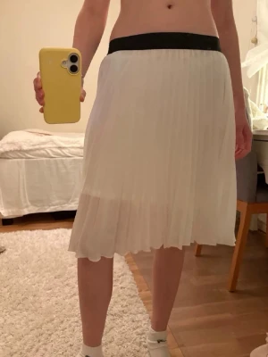 Vit plisserad kjol från Voyelles - Säljer en vit plisserad kjol från Voyelles i storlek S. Kjolen har ett svart elastiskt midjeband och är gjord i 100% polyester. Den har en luftig känsla och slutar strax ovanför knäna. Hör av dig om du undrar mått eller annat så löser jag det. Använd någon enstaka gång. 