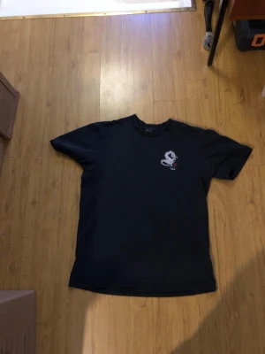 Svart t-shirt från stussy - Svart t-shirt med korta ärmar och coolt drakmotiv i vitt på ryggen samt en mindre drake på bröstet. T-shirten har en klassisk passform och är tillverkad i mjuk bomull, perfekt för en avslappnad streetwear-look. Priset är inte bestämt.