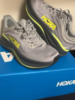 Gråa Hoka sneakers med neongula detaljer - Snygga grå sneakers från Hoka med neongula loggor och detaljer. Ovandelen är i mesh för bra andningsförmåga och sulan är tjock och svart med limegrön kant. Perfekta för dig som gillar sportig stil och vill ha bekväma skor till vardags.