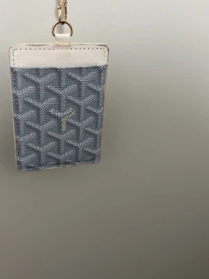 Grå korthållare med snöre - Snygg grå korthållare från  med klassiskt mönster och vita detaljer. Kommer med ett matchande snöre och guldfärgad hake, perfekt för att bära runt halsen. Tillverkad i canvas och läder, har fack både fram och bak för kort eller ID.