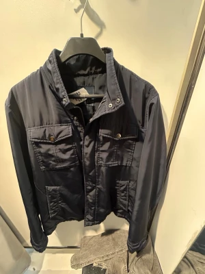 Mörkblå vindjacka med fickor - Stilren mörkblå vindjacka med hög krage och fyra framfickor med lock och tryckknappar. Jackan har dragkedja och knappstängning framtill samt raka ärmar. Perfekt för blåsiga dagar och enkel att matcha till olika outfits.