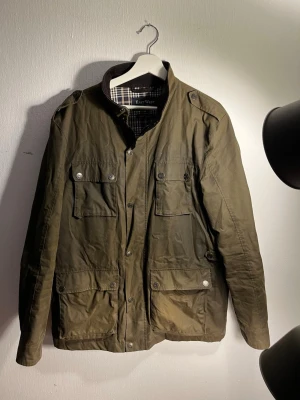 Olivgrön jacka från East West - Snygg olivgrön jacka från East West med rutigt innerfoder och fyra stora fickor framtill. Jackan har knappar, axelklaffar och en mörkbrun krage i manchester. Perfekt för dig som gillar klassisk stil med moderna detaljer.