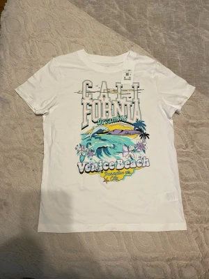 Vit t-shirt med Venice Beach-tryck - Vit t-shirt från Cubus med färgglatt tryck av texten 'California Dreaming Venice Beach' och motiv av strand, vågor och blommor. Klassisk passform med rund hals och korta ärmar. Perfekt för en avslappnad och cool stil.