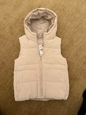 Beige dunväst med huva från Mango - Stilren beige dunväst från Mango med dragkedja framtill och två stora fickor. Västen har en mysig huva och quiltad design som ger en modern look. Perfekt för lager-på-lager och enkel att matcha med olika outfits.