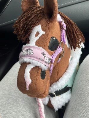 Brun käpphäst med lila grimma - Säljer en brun käpphäst med vita fläckar och man av brunt garn. Hästen har en lila grimma med tecknade hästar på och detaljer i metall. Pälsen är mjuk och det finns vita fluffiga partier runt mulen och halsen. Perfekt för lek och fantasi.