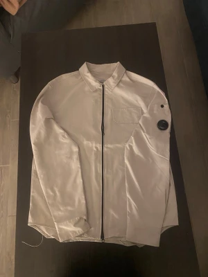 Beige overshirt från C.P. Company - Stilren beige overshirt från C.P. Company med svart dragkedja framtill, klassisk krage och bröstficka. Ärmen har den ikoniska linsen med logga. Tillverkad i ett lätt, glansigt material som ger en modern look. Perfekt för lager-på-lager och streetwear.