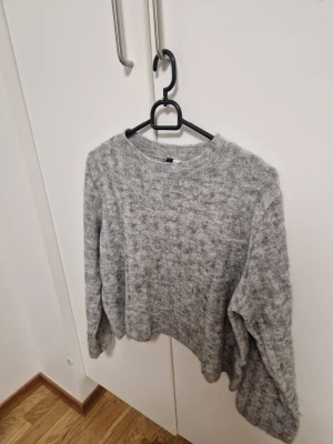 Grå stickad tröja med lös passform - Mysig grå stickad tröja med rund halsringning och avslappnad, lite boxig passform. Tröjan har långa ärmar och ribbade muddar vid hals, ärmslut och nederkant. Perfekt för lager på lager under kyliga dagar.