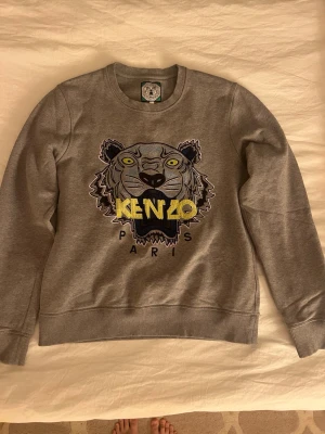 Grå Kenzo sweatshirt med tigertryck - Säljer en grå sweatshirt från Kenzo med det ikoniska broderade tigerhuvudet och texten 'KENZO PARIS' i gult och svart framtill. Tröjan har rund halsringning, ribbade muddar och är tillverkad i mjukt bomullsmaterial. I väldigt bra skick🩷