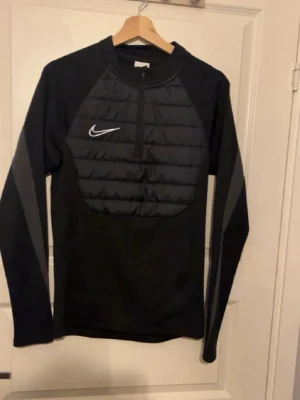 Svart långärmad träningströja Nike - Snygg svart långärmad träningströja från Nike med vadderad frontpanel och halv dragkedja. Tröjan har gråa detaljer längs ärmarna och en vit Nike-logga på bröstet. Perfekt för löpning eller träning utomhus.