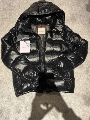 Svart pufferjacka från Moncler - Snygg svart pufferjacka från Moncler med glansig finish och huva. Jackan har dragkedja framtill, två sidofickor och klassisk Moncler-logga på ärmen. Perfekt för kalla vinterdagar och riktigt trendig look.