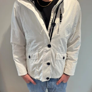Vit vindjacka från Sebago - Fräsch vit vindjacka från Sebago med broderad logga och flagga på ärmen. Jackan har stora knappar framtill, dragkedja, flera fickor och en clean look. Perfekt för dig som gillar stilren och sportig vibe. Materialet är lätt och känns skönt mot kroppen. Inga defekter och det är bara höra av er vid frågor