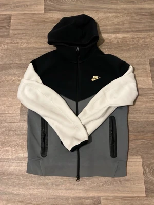 Nike hoodie svart/grå/vit - Snygg hoodie från Nike i färgblockad design med svart huva och överdel, grå kropp och vita ärmar. Dragkedja framtill, två fickor med dragkedja och Nike-logga på bröstet. Perfekt för en sportig och avslappnad stil.