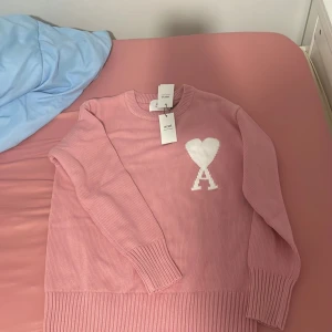 Rosa stickad tröja - Superfin rosa stickad tröja från Ami Paris med vitt hjärta och A på bröstet. Tröjan har rund halsringning, långa ärmar och breda ribbade muddar. Perfekt för dig som vill ha en mysig och trendig look.