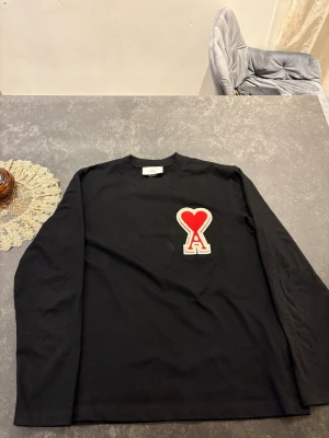 Svart långärmad topp från Ami Paris - Cool svart långärmad topp från Ami Paris med stort rött hjärta och bokstaven A broderat på bröstet. Tillverkad i mjuk bomull och har en relaxed fit. Perfekt för dig som gillar statement-plagg och vill sticka ut lite extra.