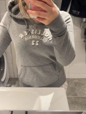 Grå Hollister hoodie med tryck - Säljer en grå hoodie från Hollister med vit broderad text 'Hollister California 22' på bröstet. Tröjan har huva med dragsko och en stor magficka framtill. Perfekt för en avslappnad och stilren look. Hoodien är i storlek M men passar mer som storlek S.