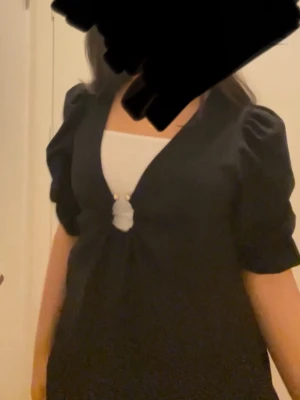 Svart blus med puffärm och ringdetalj - Trendig svart blus med djup V-ringning och snygg ringdetalj framtill. Blusen har puffiga trekvartsärmar med resår vid ärmslut och är tillverkad i ett mjukt tyg som ger en skön känsla. Perfekt för dig som gillar stilrena och moderna plagg.