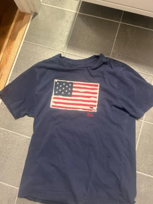  Polo Ralph Lauren t-shirt - Mörkblå t-shirt från Polo Ralph Lauren med amerikansk flagga tryckt på bröstet och RL-broderi i rött. Klassisk passform och rund hals. Perfekt för dig som vill ha en stilren och ikonisk look.