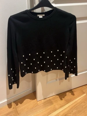 Svart tröja med pärlor - Snygg svart tröja från H&M med långa ärmar och dekorativa vita pärlor längs nederkanten och ärmarna. Tröjan har rund halsringning och är tillverkad i ett mjukt stickat material. Perfekt för en trendig look.