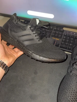 Svarta Adidas Ultra Boost sneakers - Helvita Adidas Ultra Boost sneakers i svart med stickad ovandel och svart Boost-sula. Skorna har klassiska tre ränder på sidan, snörning och en sportig, modern look. Perfekta för dig som gillar stilrena och bekväma sneakers med mycket streetkänsla.